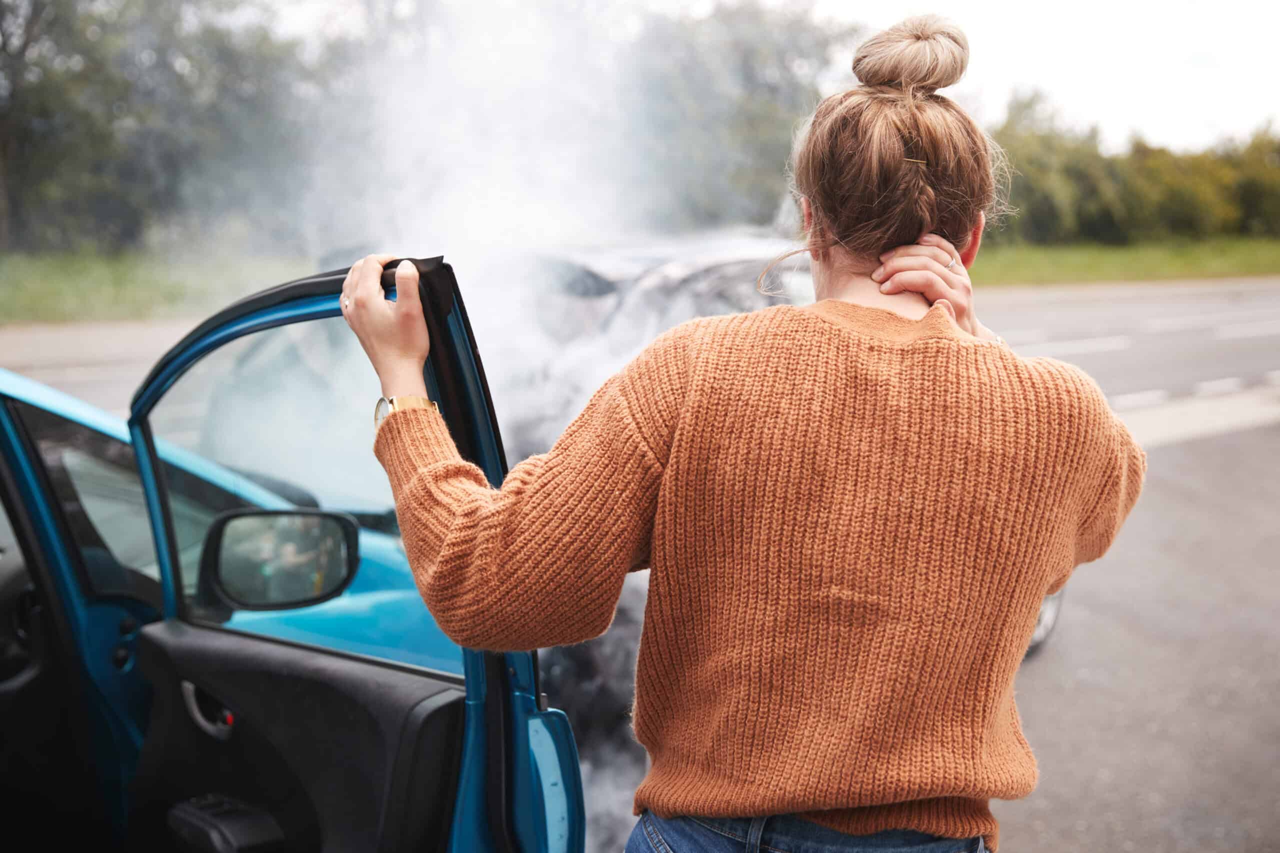 Rear view of a driver standing beside a damaged car after an accident, holding the open door and touching their neck while smoke rises from the vehicle on the roadside.