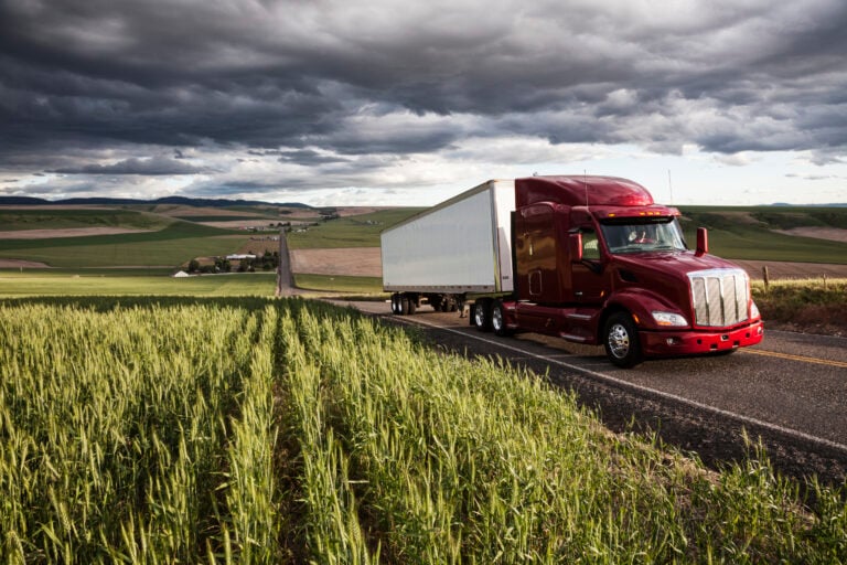 red semi truck driving on rural highway through green fields under dark storm clouds