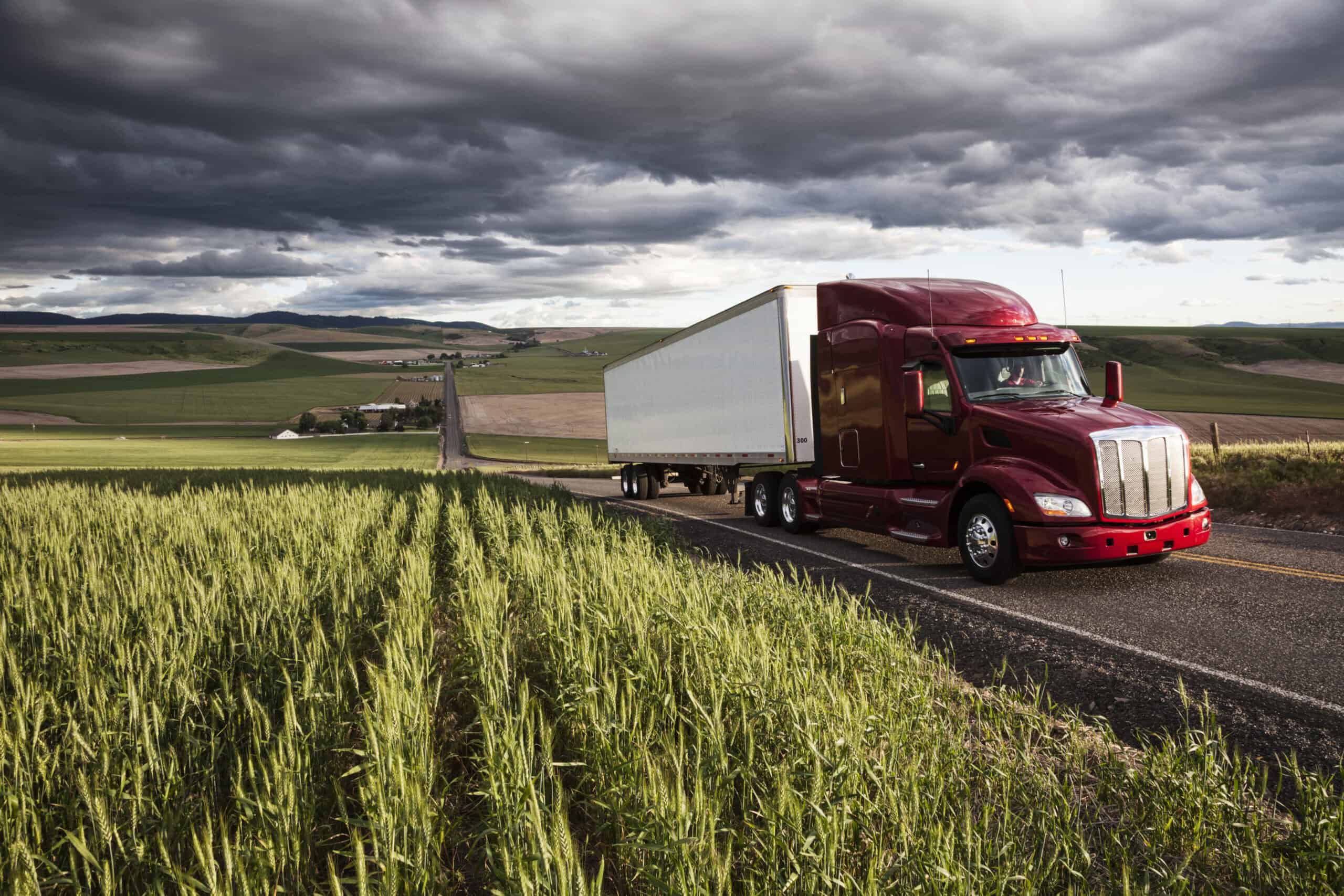 red semi truck driving on rural highway through green fields under dark storm clouds