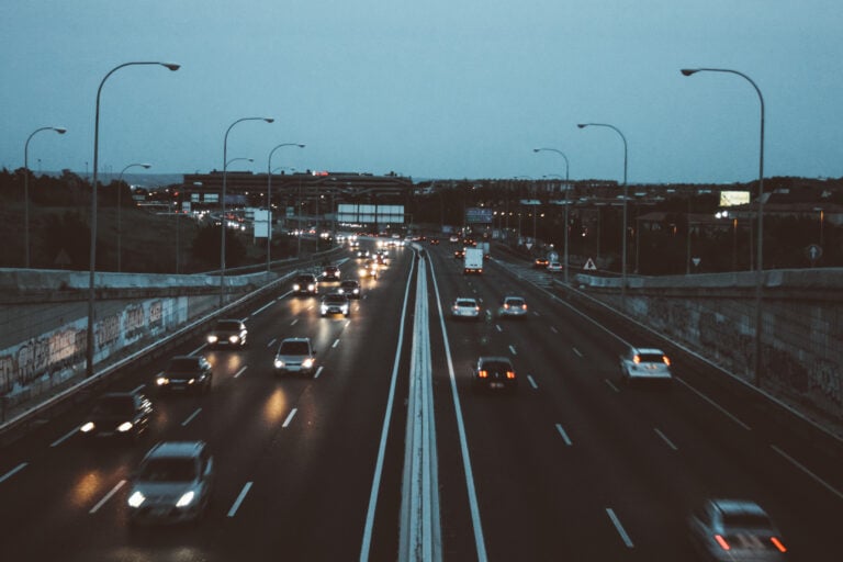 Traffic flows along a divided multi-lane highway at dusk, with headlights reflecting on the dark pavement beneath rows of curved streetlights. Buildings and road signs appear in the distance under a blue-gray sky