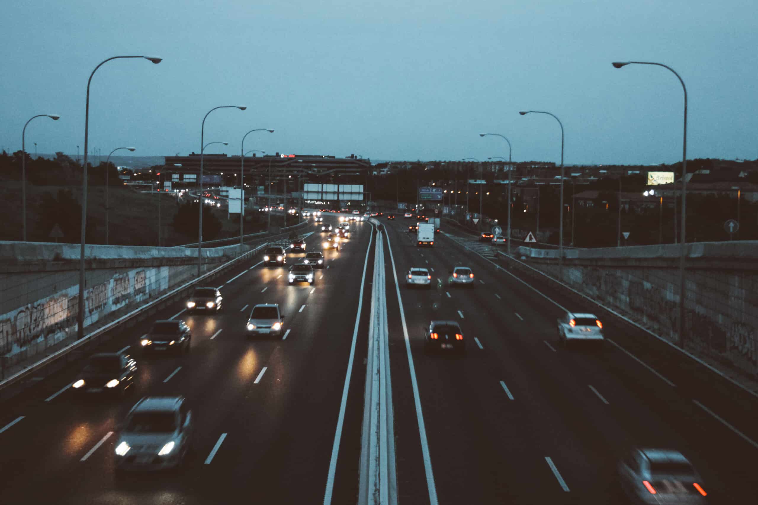 Traffic flows along a divided multi-lane highway at dusk, with headlights reflecting on the dark pavement beneath rows of curved streetlights. Buildings and road signs appear in the distance under a blue-gray sky